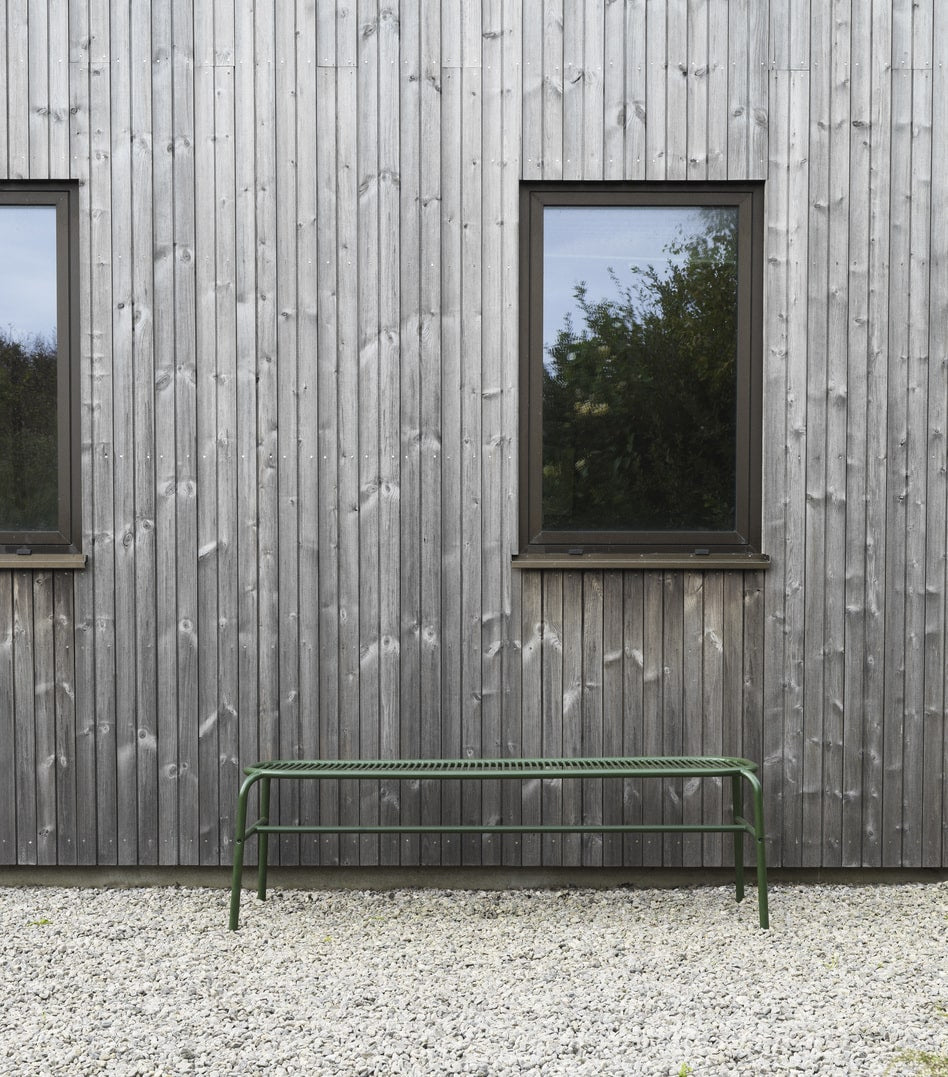 Grøn lang bænk i metal, foran hus med store vinduer