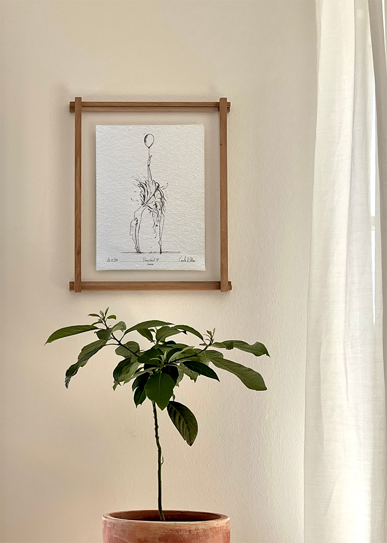 En træramme med en sort/hvid tegning af en ballerina på en hvid væg. Under billedet står der en grøn plante.