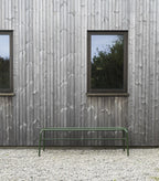 Grøn lang bænk i metal, foran hus med store vinduer 