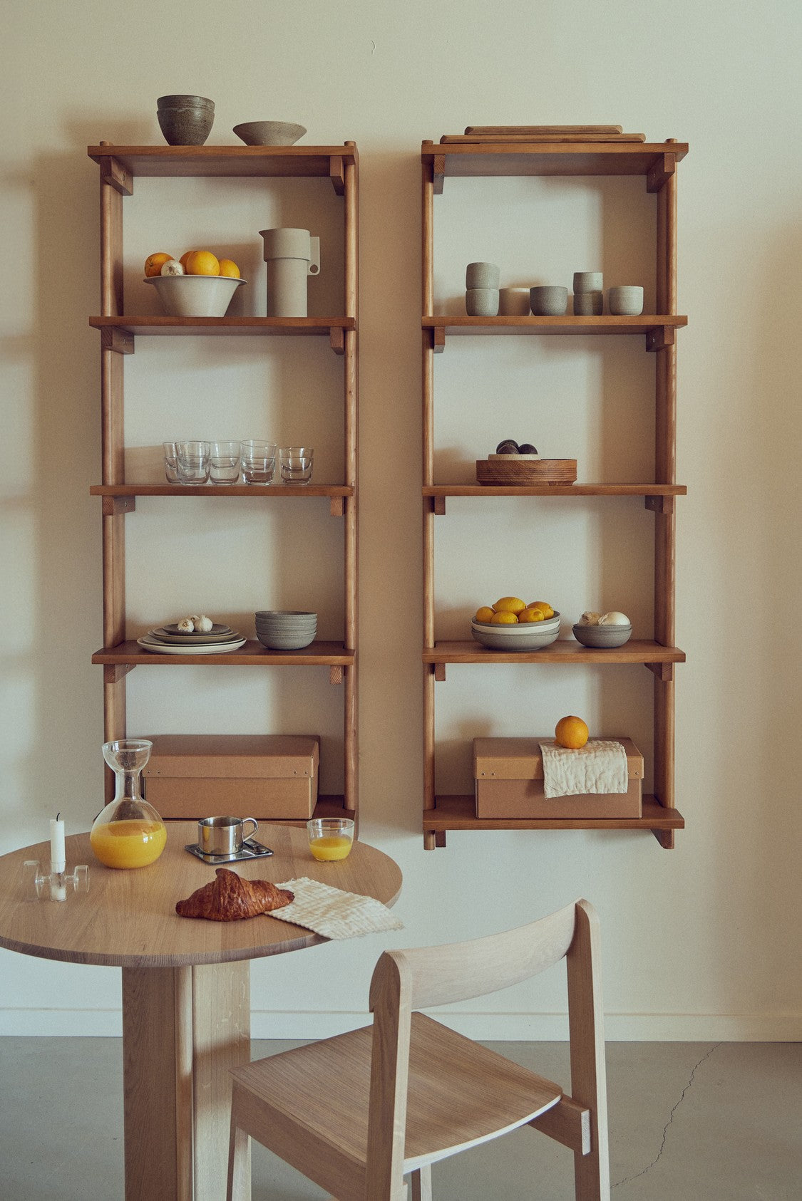 2 hylder i mørkt olieret træ med et rundt bord og en stol foran.