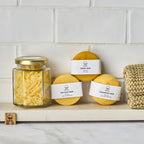 En hvid baggrund af klinker, med tre sæber, shampoo, balsam og sæbe, ved siden af står der et glas med sæbespåner. 