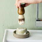 Keramisk skål med en sæbebar, mens en hånd holder en barberbørste over.