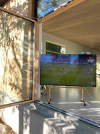 Billedet er taget udenfor, hvor man har rullet TV'et helt hen til havedøren, så man kan se fodbold udefra. TV'et hænger på en træ stander med hjul. 