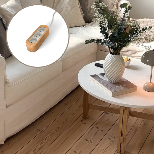 Et sofabord med hvid bordplade og træben, foran en hvid sofa. På bordet stor en lampe og en buket blomster. Et billede af en stikdåse i træ. 