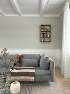 Lys og hyggelig stue med blomster på sofabordet, en grå sofa og en pyntehylde i træ monteret på væggen.