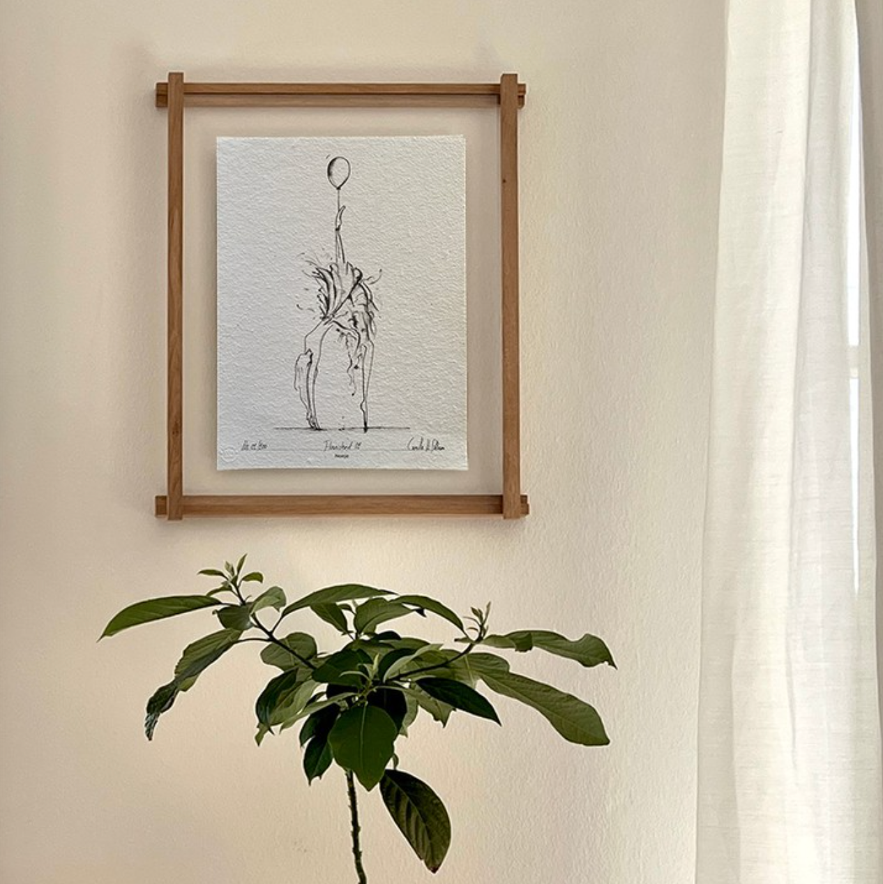 En træramme med en sort/hvid tegning af en ballerina med en ballon bundet rundt om anklen, på en hvid væg. Nedunder står der en grøn plante. 