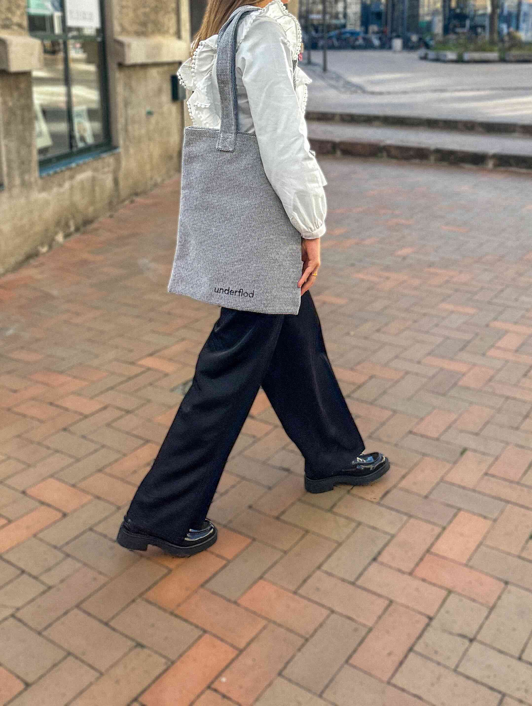 Kvinde som går udenfor på fliser med en grå mulepose, hvor der står underflod med sorte bogstaver nedst på tasken. 