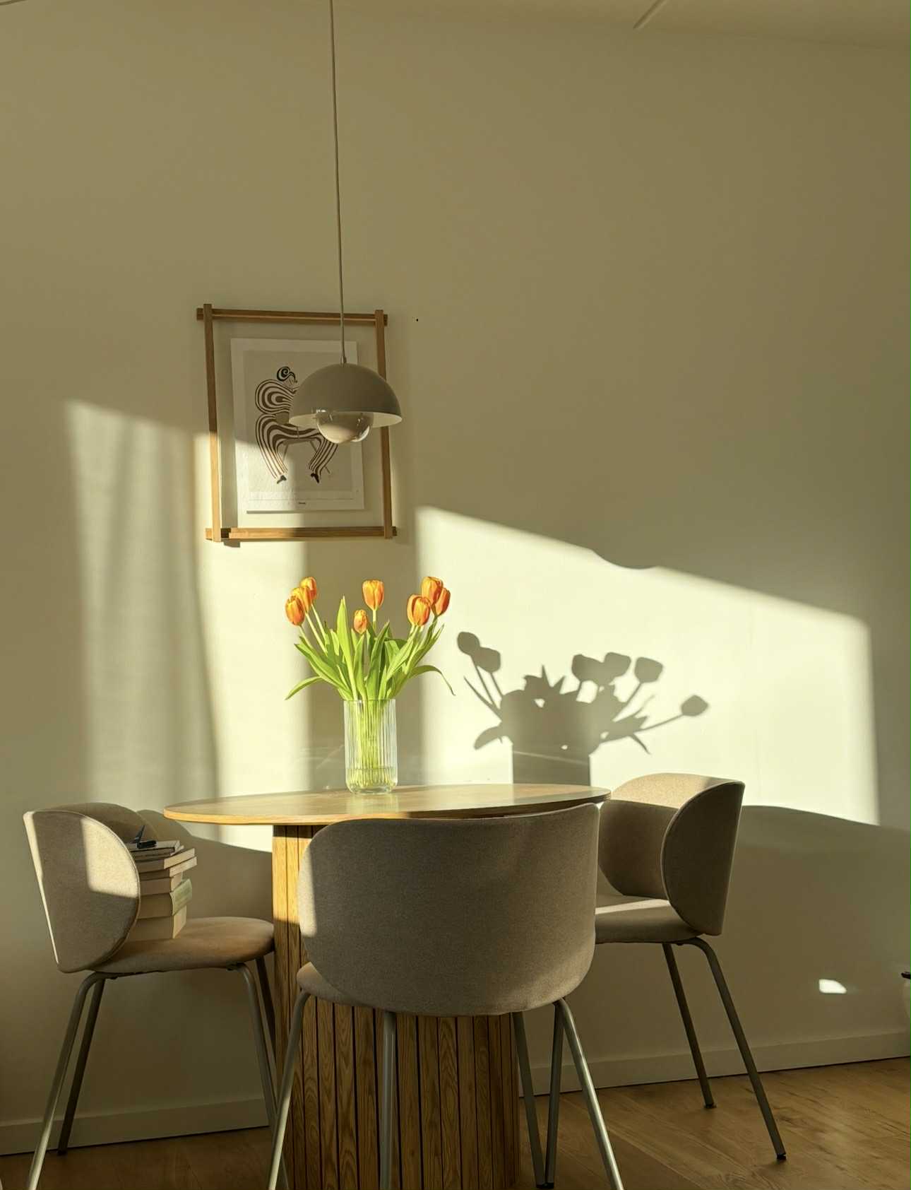 En spisestue med et bord, tre stole og en buket tulipaner. På væggen hænger en træramme på væggen med et kunsttryk af en abstrakt dame. 