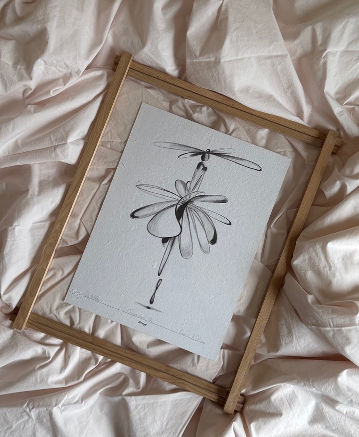 Et træramme med et kunsttryk af en sort/hvid stregtegning. Rammen ligger på noget lyserødt sengetøj. 