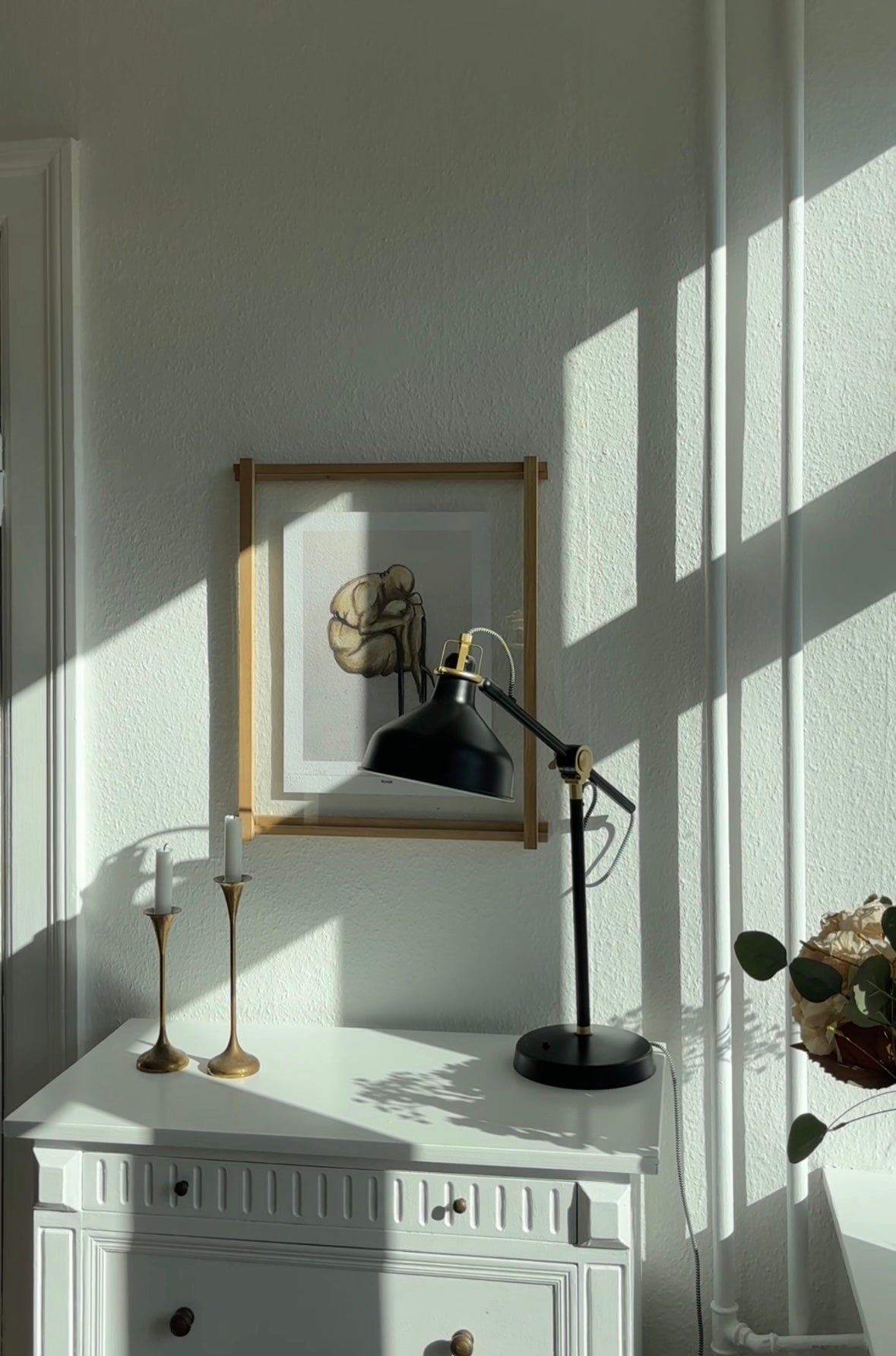 En hvid væg med et kunsttryk i en træramme, som hænger over et hvidt møbel. 